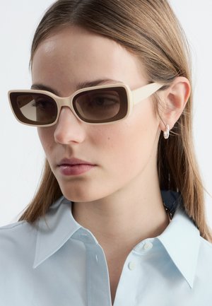 Jeune femme aux cheveux lisses châtain clair porte des lunettes de soleil rectangulaires beiges et une boucle d’oreille argentée en forme d’anneau, vêtue d’une chemise bleu clair à col.