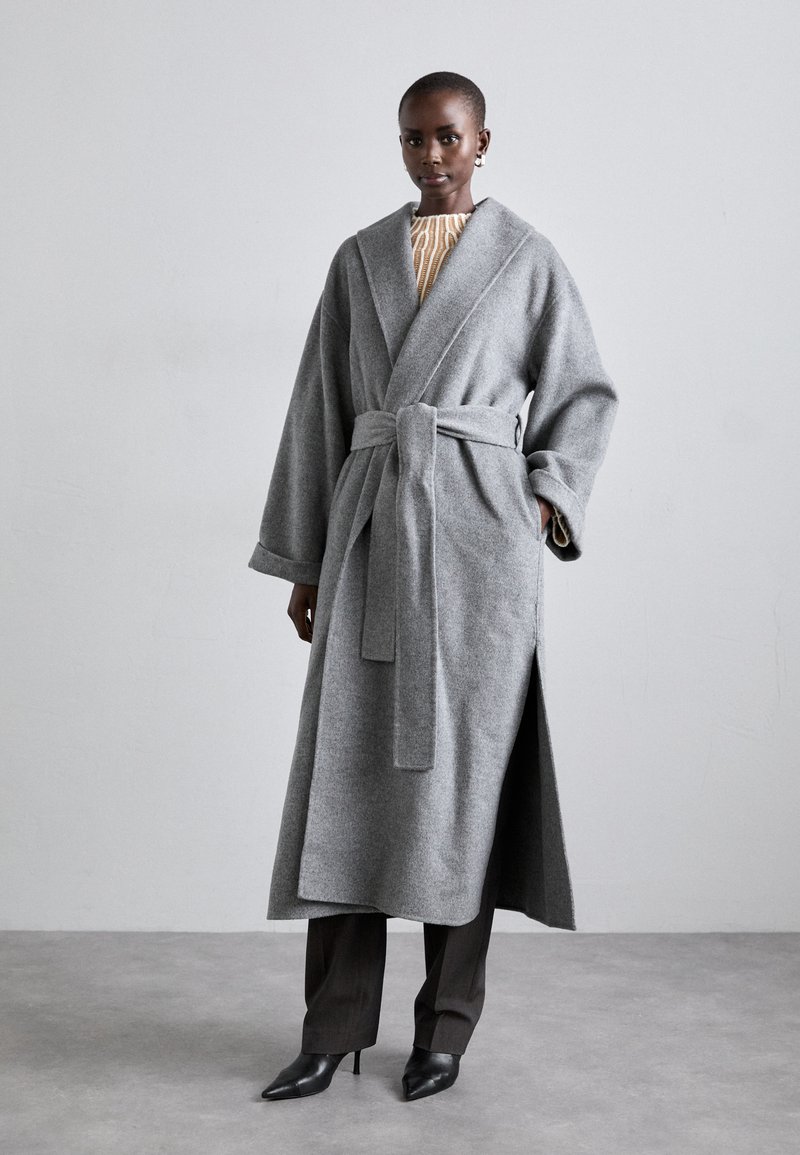 Cappotto avvolgente grigio in lana con maniche ampie, vita cinta da una cintura e spacchi laterali, abbinato a pantaloni scuri e tacchi neri a punta. Tessuto texturizzato.