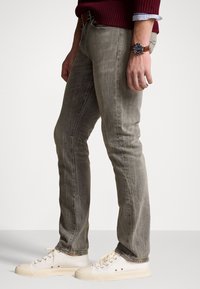 Ljusgrå smalpassande jeans med urtvättad textur och sömmar, bärs med vita sneakers och en vinröd tröja. Accenterad med en armbandsur.