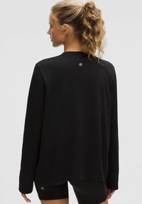 Schwarzes Langarmshirt mit lockerer Passform und strukturiertem Stoff. Verfügt über ein kleines Logo auf der Rückseite. Kombiniert mit dunklen Shorts.