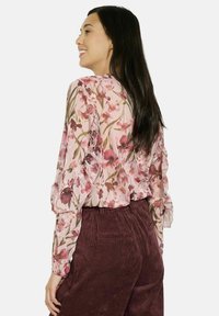 Ziedu blouse ar rozā fona, kurai ir viļņotas piedurknes un zaļie akcenti. Apvienota ar teksturētām bordo velveta biksēm.