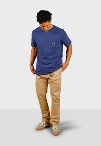 Blå bomulls-t-shirt med en ficka som har en grå logotyp, ihopparad med beige byxor och grå sneakers med vita snörningar och orange sulor.