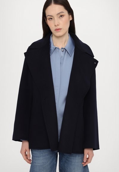 Cappotto in misto lana blu navy con ampio risvolto, design aperto e maniche oversize. Indossato sopra una camicia button-up azzurro chiaro e jeans.