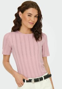 Top corto a maniche corte di colore rosa chiaro, realizzato in maglia, con texture a coste, scollatura ondulata e silhouette aderente, indossato con una cintura nera chiodata.