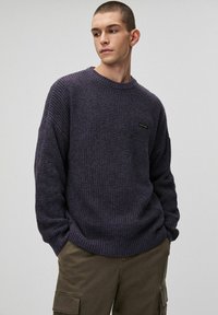 Oversized fioletowy sweter z dzianiny o teksturowanej tkaninie, z prążkowanymi mankietami i dołem, z małym czarnym logo na piersi.