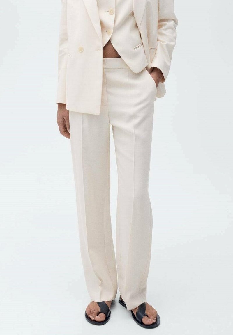 Personne portant un costume crème avec un blazer court, un pantalon taille haute à jambes larges et des sandales noires à bouts ouverts sur fond blanc.