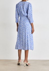 Modré midi šaty se smockovaným pasem, tříčtvrtečními rukávy a bílými abstraktními vzory; spárované se světlými otevřenými podpatky.