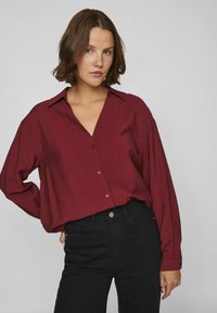 Camisa burdeos de manga larga con escote en V, botones frontales y corte relajado, combinada con jeans negros, sobre un fondo gris.