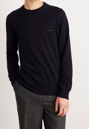 Mężczyzna ubrany w czarny sweter z długim rękawem z logo „BOSS” oraz szare spodnie w prążki, stojący na tle jasnego, jednolitego tła.