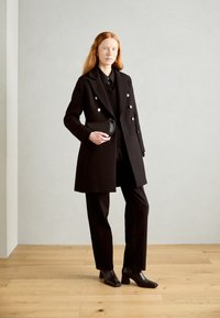 Abrigo negro de doble botonadura con botones plateados, superpuesto a un suéter y pantalones negros, combinado con botas de tacón negras y un bolso pequeño.