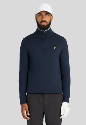 Mand iført marineblå quarter-zip sweater, hvid perforeret kasket, sorte bukser og hvid golfhandske, stående mod ensfarvet baggrund.