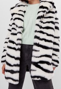 Kabát z umělé kožešiny s černobílými pruhy zebry, širokým límcem, velkými knoflíky a měkkou, plyšovou texturou. Nosit na světle zeleném svetru.