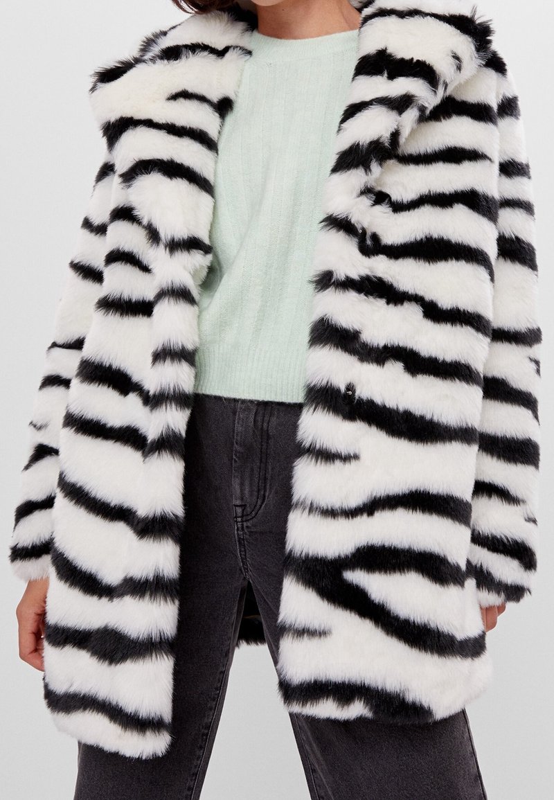 Kabát z umělé kožešiny s černobílými pruhy zebry, širokým límcem, velkými knoflíky a měkkou, plyšovou texturou. Nosit na světle zeleném svetru.