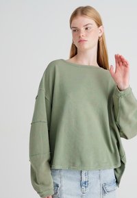 Olivengrønn, stripet langermet topp med løs passform, vid halslinning og senket skulder, kombinert med lyse denimshorts.