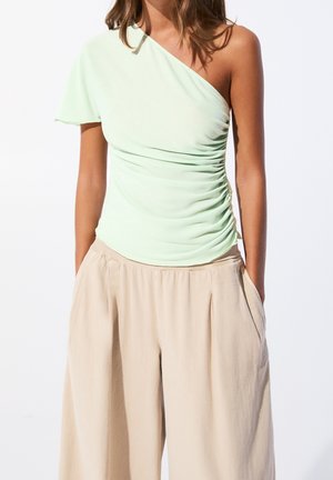 Femme portant un haut vert clair asymétrique froncé et un pantalon beige ample, les mains dans les poches.