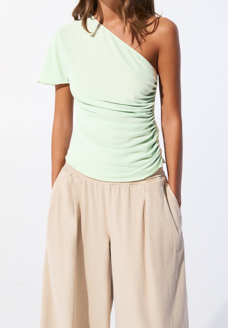 Femme portant un haut vert clair asymétrique froncé et un pantalon beige ample, les mains dans les poches.