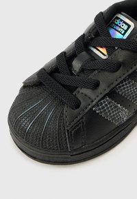 Mustad toss ülemise tekstureeritud nahaga, millel on kummist tald, mustad lamedad paelad ja kolm tumedat triipu. Keele peal helkiv logo.