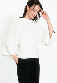 Blusa bianca con maniche larghe e fluide, realizzata in un tessuto morbido. Abbinata a pantaloni neri, caratterizzati da una vestibilità comoda e tasche laterali.