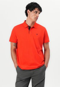 Rød bomulds polo shirt med sort krave og et logo på brystet; korte ærmer og en afslappet pasform. Iført med grå bukser.