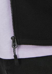 Black fleece jacket with a zipper and a pull tab, layered over a light purple shirt, featuring visible stitching along the hem.