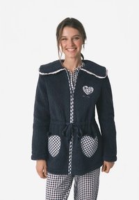 Laivaston sininen fleece-takki, jossa suuri kaulus, valkoinen reunus ja sydänkuvio. Takissa on ruutukuviolliset taskut ja vyötäröllä solmittava vyö.