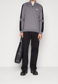 Mann in grauem Quarter-Zip-Pullover mit schwarzen Streifen, schwarzen Hosen, schwarzen Stiefeln und einer schwarzen Ledertasche an seiner Seite.