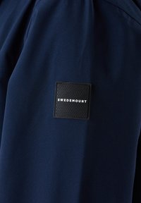 Giacca di tessuto blu navy con una toppa nera quadrata etichettata "SWEDEMOUNT" caratterizzata da una superficie testurizzata. Il design è semplice e privo di decorazioni.