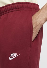 Maroon sweatpants with a white Nike logo. Features an elastic waistband and side pockets. Material appears soft with a smooth texture.