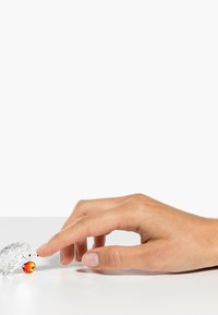 Figura de erizo de cristal con un acento naranja facetado, colocada sobre una superficie plana, con una mano humana extendiéndose hacia ella.