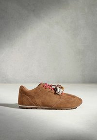 Baskets en daim marron avec des lacets rouges et des breloques florales décoratives, sur un fond gris uni.