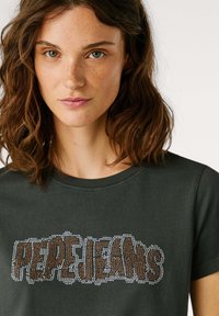 Camiseta de manga corta en verde oscuro con "PEPE JEANS" en brillantes cristales marrones y blancos, escote redondo y material de algodón suave.