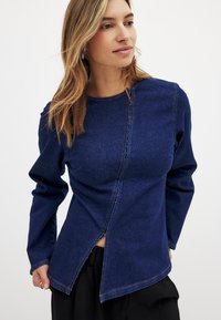 Tumši zils džinsi auduma garām piedurknēm tops ar unikālu diagonālu šuvi un šķeltu priekšu. Iespējams, ar kontrastējošiem oranžiem diegiem un apaļu izgriezumu.