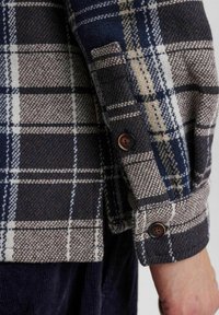 Plaid shirt made of wool blend fabric; features navy, cream, and brown colors; two buttons on the cuff; soft, textured finish.