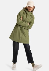 Olive green waterproof jacket with a hood, front zipper, and side pockets, paired with dark jeans and light-colored sneakers.