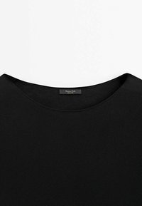 Haut en maille noir à col rond avec une petite étiquette en tissu cousue à l’intérieur du col indiquant la marque et la taille.