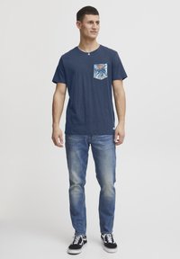 T-paita tummansinisellä värillä, jossa on kuusikulmainen tasku trooppisella printillä, yhdistettynä vaaleansinihin farkkuihin ja mustiin lenkkareihin.