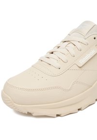 Jasnobeżowy sneaker marki Reebok wykonany z teksturowanej skóry, posiadający okrągły nosek, perforacje, sznurowadła oraz biały logo po boku.