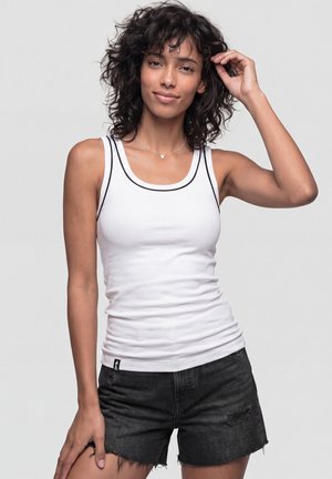 Junge Frau mit lockigem Haar, die ein weißes Tanktop und schwarze, abgenutzte Shorts trägt, lächelt mit einer Hand in der Nähe ihrer Haare.