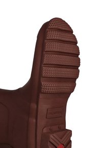 Suela de bota de goma marrón con patrones de texturización, surcos para tracción y un pequeño detalle en rojo cerca del talón.