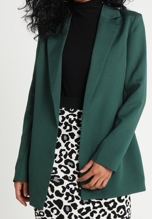 Femme portant un blazer vert foncé sur un haut noir avec une jupe à imprimé léopard noir et blanc, mains détendues sur les côtés.