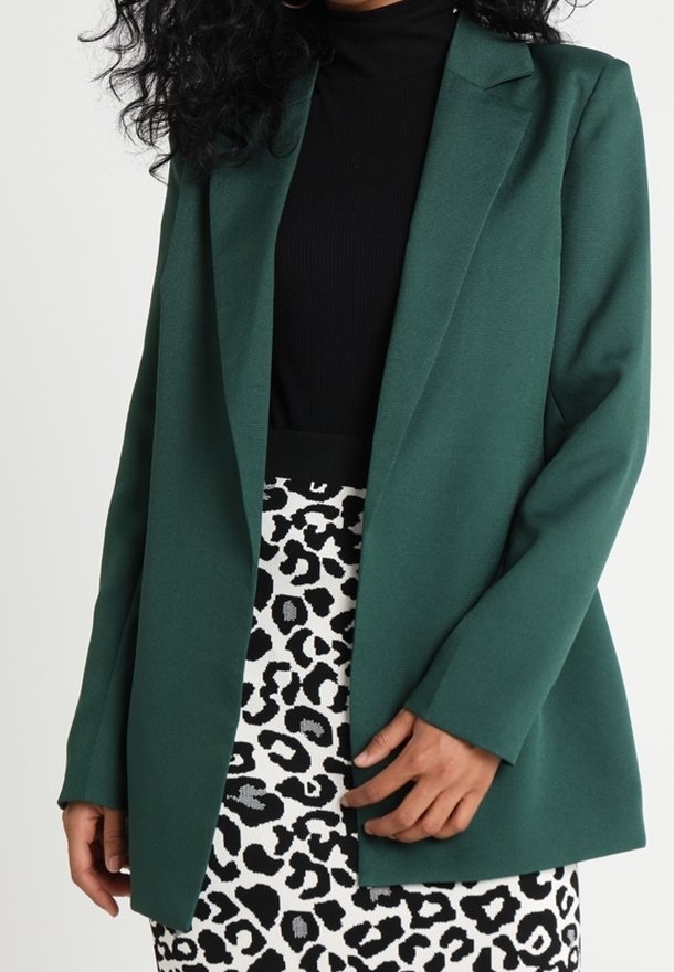 Femme portant un blazer vert foncé sur un haut noir avec une jupe à imprimé léopard noir et blanc, mains détendues sur les côtés.