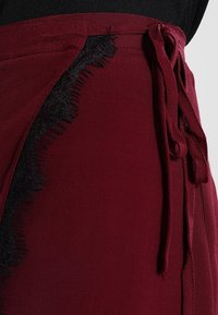 Gonna avvolgente bordeaux con un accento in pizzo nero texturizzato. Presenta una vita con fiocco e dettagli sfrangiati lungo i bordi. Finitura in tessuto liscia.