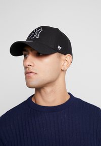 Sort baseballcap med frontlogo, buet skygge og sømdetaljer. Båret med en marineblå ribbestrikket genser. Diskré brodert merkevarelogo på siden.