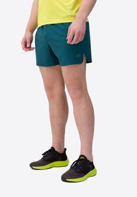 Teal sportshorts met zijsplitten, een logo op het linkerbeen, gecombineerd met zwart en neon groene sneakers met gestructureerde zolen.
