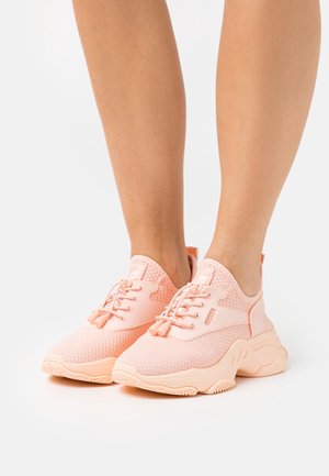 Personne portant des baskets en tissu tricoté rose clair avec des semelles épaisses et des fermetures à lacets, debout sur un fond blanc.