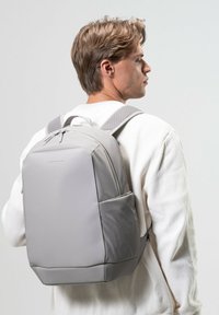 Grå ryggsäck tillverkad av slät material, med vadderade remmar, en strukturerad form och en sidoficka. Enkel design utan synliga mönster.