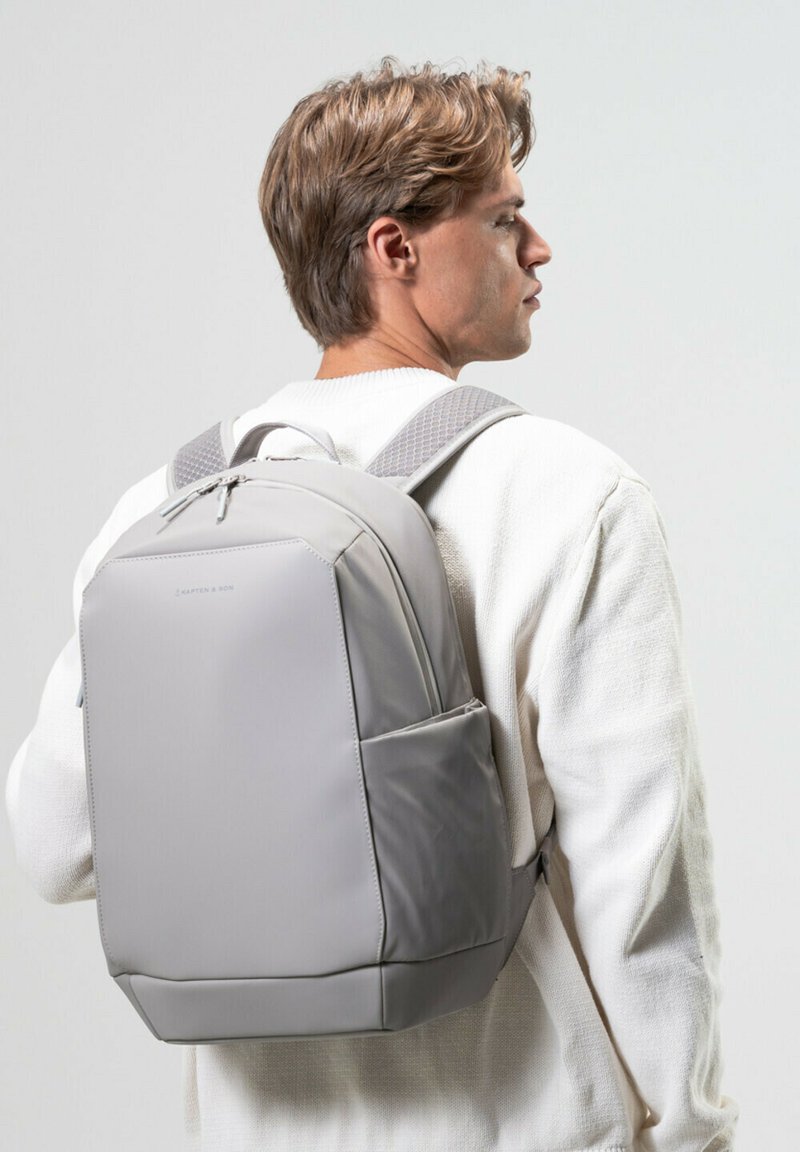 Grå ryggsäck tillverkad av slät material, med vadderade remmar, en strukturerad form och en sidoficka. Enkel design utan synliga mönster.