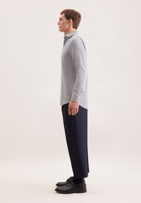 Uomo in profilo che indossa una camicia a maniche lunghe a quadri, pantaloni navy alla caviglia, calzini neri e scarpe eleganti nere.