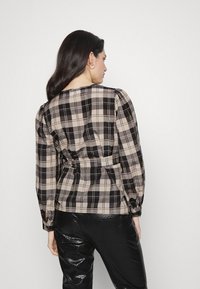 Blusa xadrez em bege e preto, com mangas bufantes e cintura ajustada. Usada com calças de couro sintético pretas e brilhantes.