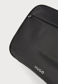 Melnas auduma soma ar gludu tekstūru, ar rāvējslēdzēja aizdari un smalku reljefu logo "HUGO" baltā krāsā. Minimālisma dizains ar noapaļotiem stūriem.